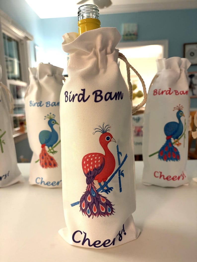 May include: White fabric wine bottle bags with a drawstring closure. Each bag features a colourful peacock illustration and the words "Bird Bam" and "Cheers!". A yellow bottle is visible in the top of one bag.