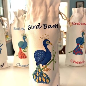 May include: White fabric wine bottle bags with a drawstring closure. Each bag features a blue peacock illustration with a yellow and orange tail, bamboo stalks, and the words "Bird Bam" and "Cheers!" in blue script.