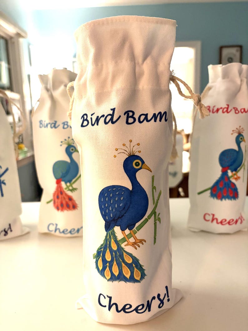 May include: White fabric wine bottle bags with a drawstring closure. Each bag features a blue peacock illustration with a yellow and orange tail, bamboo stalks, and the words "Bird Bam" and "Cheers!" in blue script.