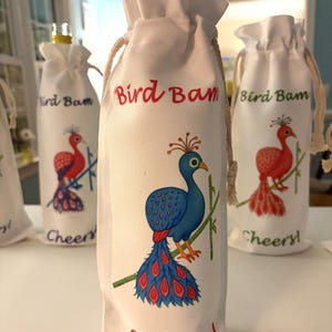 May include: White drawstring wine bottle bags featuring a peacock illustration in blue, red, and green. The text "Bird Bam" and "Cheers!" are printed on the bags. The bags are made of a soft fabric.