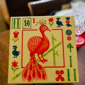 May include: Square, cream-coloured tile featuring a red bird with a long tail, perched on green bamboo stalks. The tile has a red border and is decorated with various symbols and Chinese characters. The tile is displayed on a black metal stand.