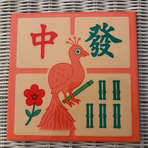 May include: Square tile with a coral border featuring a stylized pink bird holding a green bamboo stalk. The tile has Chinese characters and a red flower. The background is a light beige colour.