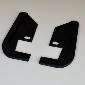 May include: Two black, L-shaped brackets with rounded corners and a small oval hole. The brackets are made of a matte material and are positioned against a white background.