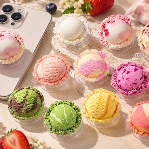 May include: A collection of round, ice cream-shaped phone grips featuring various colors and textures, including pink, green, yellow, and white scoops with textured edges, one attached to a light-colored smartphone, arranged on a flat surface.