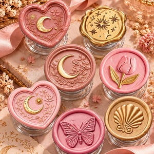 May include: A collection of seven wax seal stamps featuring various shapes and designs. The stamps include heart and circular forms in shades of pink and gold, displaying embossed motifs of a crescent moon, stars, a tulip, and a butterfly.