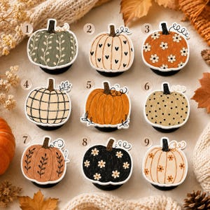 May include: A collection of nine circular phone grips featuring pumpkin illustrations numbered one through nine. Designs include sage green with vines, cream with black hearts, orange with white flowers, plaid, solid orange, polka dots, and black with flowers.
