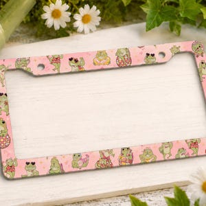 May include: A rectangular license plate frame featuring a light pink background with a repeating pattern of green frogs in various poses, including wearing heart-shaped sunglasses, sitting on disco balls, and holding small pink accessories.