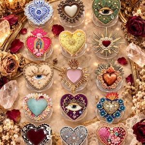 May include: A collection of fourteen heart-shaped ornaments arranged in a grid. Each piece features distinct designs including embroidered eyes, floral motifs, metallic sunburst frames, and religious imagery in various colors like blue, gold, pink, and red.