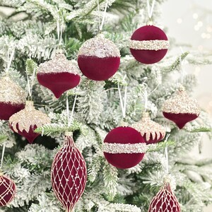 May include: Assortment of Christmas tree ornaments in various shapes and designs. The ornaments are primarily burgundy with gold glitter accents. Some are round, others teardrop-shaped, and some have a lattice pattern. White ribbons are used for hanging.