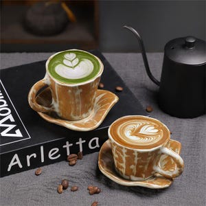 May include: Two ceramic coffee cups with matching saucers. One cup contains a green beverage with white latte art, the other a brown beverage with latte art. The cups and saucers have a textured, brown and white design. The word "Arletta" is visible on a black book.