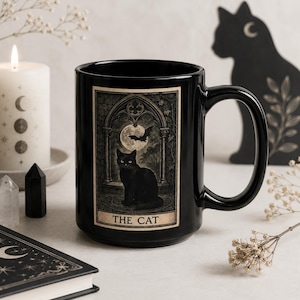 May include: A black ceramic mug featuring a rectangular graphic of a black cat sitting in front of a stone archway with a full moon and a flying bat. The text THE CAT is printed in a serif font at the bottom of the graphic within the design.