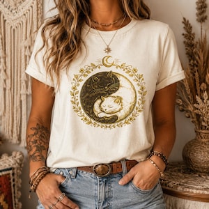 May include: A cream-coloured short-sleeve t-shirt featuring a central graphic of a black cat and a white cat curled together in a yin-yang formation. The cats are encircled by a gold-toned floral wreath with a crescent moon symbol at the top.
