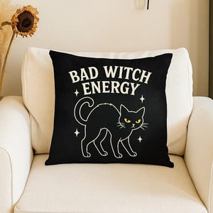 Può includere: Cuscino quadrato nero con la scritta bianca "BAD WITCH ENERGY" e un'illustrazione di un gatto nero. Il gatto ha occhi gialli e stelle bianche. Il cuscino è su una poltrona bianca.