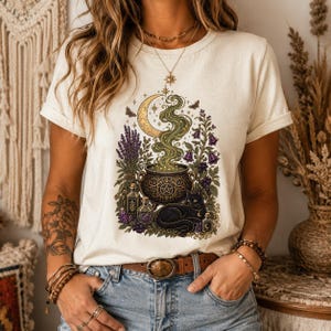 Może przedstawiać: Kremowy t-shirt z krótkim rękawem z nadrukiem przedstawiającym czarnego kota siedzącego obok kociołka z wirującym zielonym dymem, półksiężycem, gałązkami lawendy, fioletowymi dzwonkami i małymi szklanymi buteleczkami z miksturami.