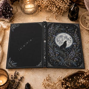 Peut inclure: Un livre à couverture rigide noire présentant une silhouette de loup argenté hurlant à la pleine lune entourée d'illustrations florales bleues sur la couverture, avec des empreintes de pattes et des volutes aux coins.