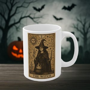 May include: White ceramic mug featuring a tarot card design with a witch illustration. The card reads "The Witch" and depicts a woman in a witch hat, holding a wand and conjuring flames. The background includes a sun, moon, and stars.