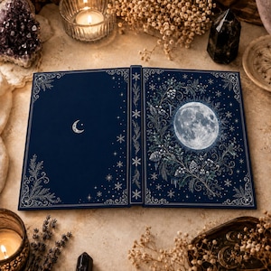 May include: A dark blue hardcover book featuring a silver crescent moon on the front cover and a full moon surrounded by holly, pinecones, and snowflakes on the back cover. The spine and edges are decorated with intricate silver snowflake patterns.