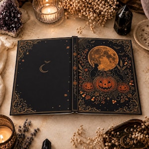 Peut inclure: Un livre à couverture rigide noir orné d'illustrations dorées représentant un croissant de lune et une chauve-souris à gauche, ainsi qu'une pleine lune, un chat et trois citrouilles entourés de feuillage d'automne et de toiles d'araignée.