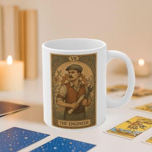 Puede incluir: Taza de cerámica blanca con un diseño de carta del tarot de "El Ingeniero". La ilustración representa a un hombre con gorra y chaleco sosteniendo una llave inglesa y planos. La taza tiene un asa curva.