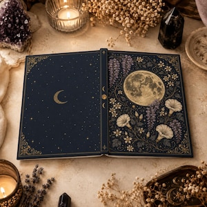 Könnte beinhalten: Ein gebundenes Buch mit dunkelblauem Einband, das links eine goldene Mondsichel mit Sternen und rechts einen Vollmond umgeben von weißen Blüten und violettem Blauregen zeigt. Der Buchrücken zeigt drei Mondphasen in Goldfolie.