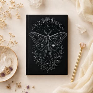 Peut inclure: Un carnet à couverture rigide noire présentant une illustration au trait blanc d'un grand papillon de nuit centrée sur la couverture. Au-dessus du papillon se trouve un arc de phases lunaires, et en dessous, des vignes florales et un croissant de lune.