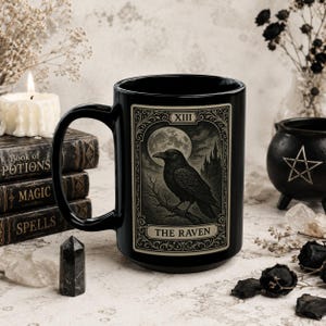 Può includere: Una tazza in ceramica nera con una grafica rettangolare disegnata come una carta dei tarocchi. L'illustrazione raffigura un corvo nero appollaiato su un ramo sotto una luna piena con un castello sullo sfondo. Il testo riporta XIII e THE RAVEN.