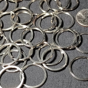 May include: A collection of silver-colored key rings, many of which are interlocked, are scattered across a dark gray surface. A U.S. quarter coin is visible in the upper right corner. The key rings are circular and made of metal.