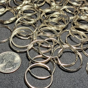 May include: A collection of shiny, gold-colored key rings, many overlapping, with a quarter coin for size comparison. The key rings are circular and appear to be made of metal. The background is a dark, textured surface.