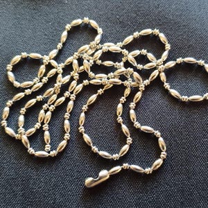 May include: A silver-colored necklace with a series of small, oval-shaped beads connected by tiny links. The necklace is lying on a dark blue textured surface. The clasp is visible at the end of the necklace.