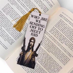 Puede incluir: Marcap&aacute;ginas con el texto "WHO ARE WE SENDING OFF TO MEET DAD" y una imagen de Jes&uacute;s sosteniendo dos ametralladoras. El marcap&aacute;ginas tiene una borla dorada y est&aacute; sobre un libro abierto.