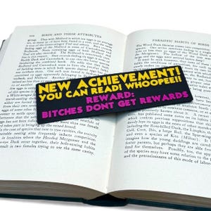 Puede incluir: Un marcap&aacute;ginas negro con texto amarillo y rosa que dice "NEW ACHIEVEMENT! YOU CAN READ! WHOOPEE!! REWARD: BITCHES DONT GET REWARDS" est&aacute; colocado en un libro abierto.