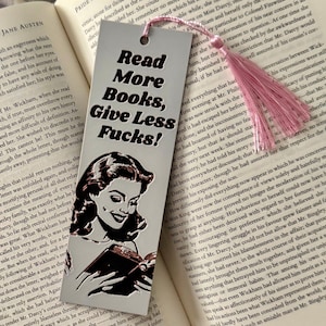 May include: A silver bookmark with a pink tassel, featuring the text "Read More Books, Give Less Fucks!" and a black and white illustration of a woman reading a book. The bookmark is placed in a book.