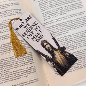 Puede incluir: Marcap&aacute;ginas con el texto "WHO ARE WE SENDING OFF TO MEET DAD" y una imagen de Jes&uacute;s con rifles. Tiene una borla dorada y est&aacute; colocado sobre p&aacute;ginas de libro abiertas.