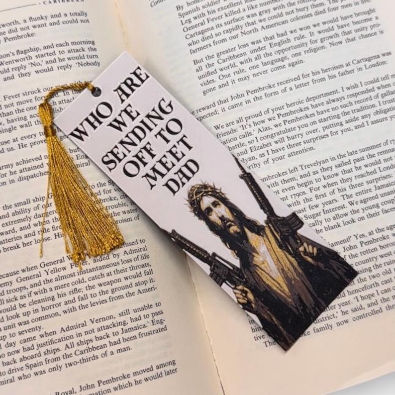 Puede incluir: Marcap&aacute;ginas con el texto "WHO ARE WE SENDING OFF TO MEET DAD" y una imagen de Jes&uacute;s con rifles. Tiene una borla dorada y est&aacute; colocado sobre p&aacute;ginas de libro abiertas.