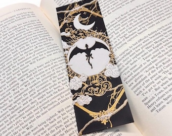 Puede incluir: Marcapáginas negro con la silueta de un dragón dentro de un círculo blanco, sobre un diseño de luna y nubes. Los detalles dorados y los elementos decorativos realzan el tema de fantasía. El marcapáginas está sobre un libro abierto.
