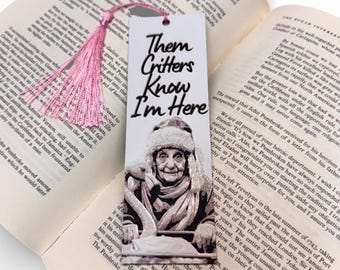 Puede incluir: Un marcapáginas blanco con el texto "Them Critters Know I'm Here" y una imagen de una persona mayor con sombrero. Una borla rosa cuelga de la parte superior. El marcapáginas está en un libro abierto.