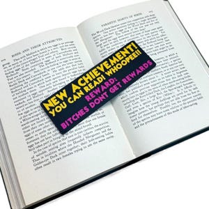 Puede incluir: Un marcap&aacute;ginas negro con texto amarillo y rosa que dice "NEW ACHIEVEMENT! YOU CAN READ! WHOOPEE!! REWARD: BITCHES DON'T GET REWARDS" est&aacute; colocado en un libro abierto. Las p&aacute;ginas del libro est&aacute;n llenas de texto.