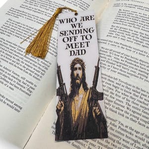 Puede incluir: Marcap&aacute;ginas con el texto "WHO ARE WE SENDING OFF TO MEET DAD" y una imagen de Jes&uacute;s sosteniendo dos rifles. El marcap&aacute;ginas tiene una borla dorada y est&aacute; sobre un libro abierto.