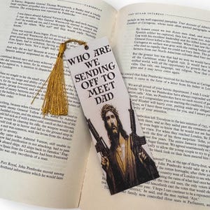 Puede incluir: Marcap&aacute;ginas con el texto "WHO ARE WE SENDING OFF TO MEET DAD" y una imagen de Jes&uacute;s sosteniendo dos rifles. El marcap&aacute;ginas tiene una borla dorada y est&aacute; sobre un libro abierto.