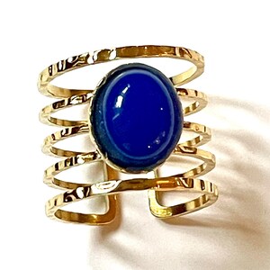 Peut inclure: Bague manchette dorée avec une grosse pierre précieuse ovale bleu foncé. La bague a une texture martelée et un design multi-bandes, créant un accessoire audacieux. La pierre précieuse est au centre, sertie dans une lunette dorée.