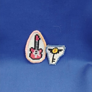 Könnte beinhalten: Zwei kleine, handgefertigte Kreuzstich-Patches. Einer hat eine ovale Form mit einem rosa Rand und einer roten und weißen Gitarre. Der andere ist ein Schlüsseldesign mit einem gelben Kreis oben und einem blauen Rand. Beide befinden sich auf blauem Hintergrund.