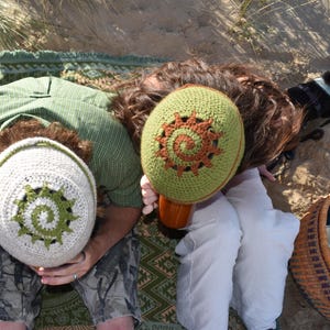 May include: Two crocheted beanies featuring a spiral sunburst design. One beanie is off-white with a green spiral sunburst, and the other is green with a brown spiral sunburst. Both hats are round, textured, and crafted from yarn.
