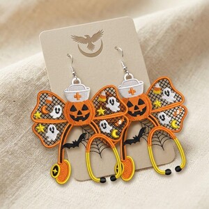Peut inclure: Une paire de boucles d'oreilles brodées représentant une citrouille d'Halloween portant une coiffe d'infirmière blanche, reliée à un stéthoscope. La citrouille orange est entourée d'une toile noire avec des fantômes, des étoiles et des chauves-souris.