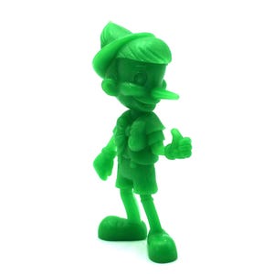 May include: A solid green figurine of Pinocchio, the classic Disney character. The figure is standing with a thumbs-up gesture, wearing a hat, shorts, and a vest. The long nose is a prominent feature. The figurine is set against a white background.