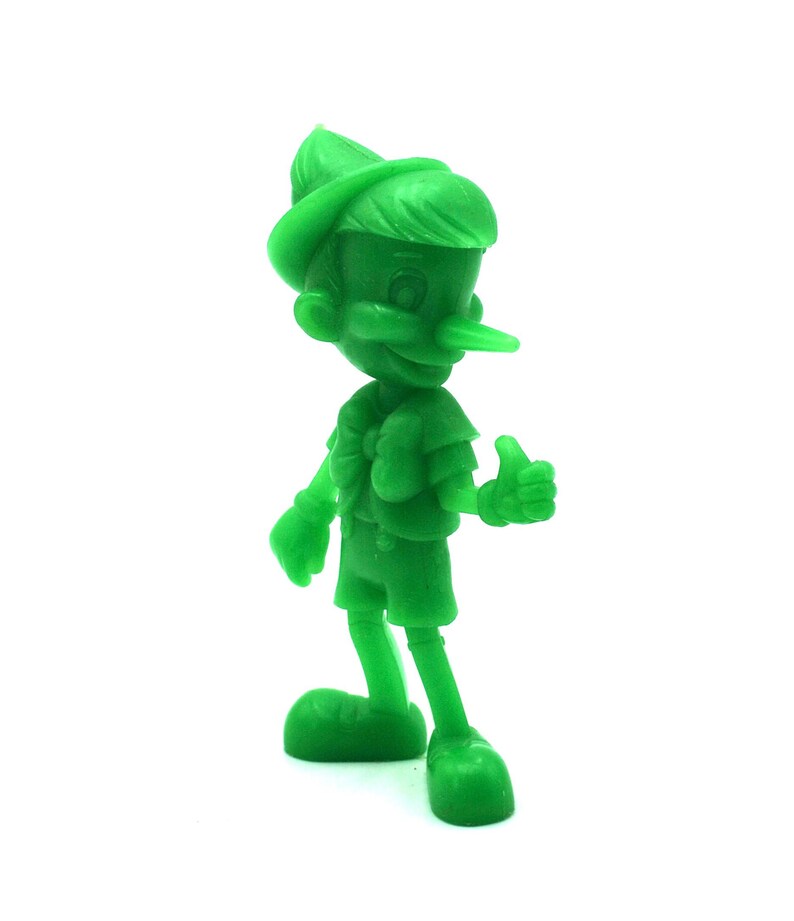 May include: A solid green figurine of Pinocchio, the classic Disney character. The figure is standing with a thumbs-up gesture, wearing a hat, shorts, and a vest. The long nose is a prominent feature. The figurine is set against a white background.