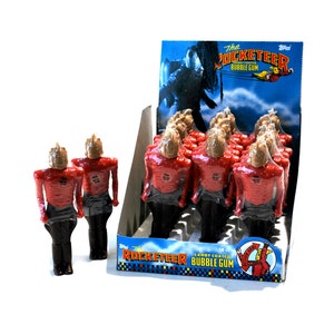 Peut inclure: Une boîte d'exposition de 18 figurines de chewing-gum enrobées de bonbons représentant The Rocketeer, un personnage de super-héros du film du même nom sorti en 1991. Les figurines sont rouges et dorées et sont emballées dans une enveloppe en plastique transparente. La boîte est bleue et présente une image de The Rocketeer volant dans les airs. Le texte sur la boîte indique "The Rocketeer Candy Coated Bubble Gum."