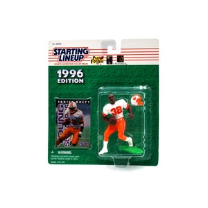 May include: A plastic action figure of a football player wearing a white and orange uniform with the number 38 on the front. The figure is holding a football and is posed in a running position. The figure is packaged in a plastic blister pack with a green background. The package also includes a card with a picture of the player and the text "1996 Edition" and "Starting Lineup".