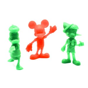 May include: Three colorful plastic figurines of cartoon characters: Donald Duck in green, Mickey Mouse in red, and Pinocchio in green. The figures are standing on a white surface.