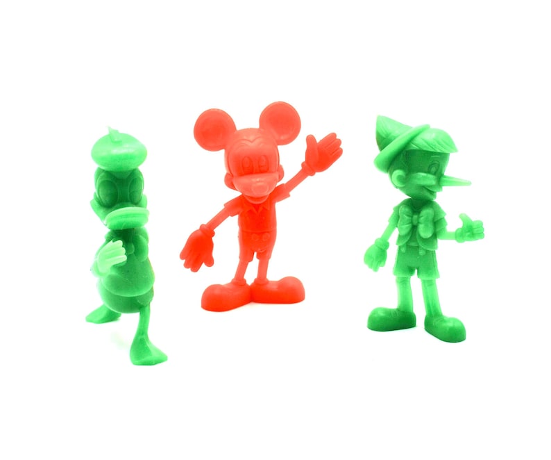 May include: Three colorful plastic figurines of cartoon characters: Donald Duck in green, Mickey Mouse in red, and Pinocchio in green. The figures are standing on a white surface.