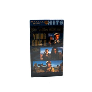May include: A VHS tape for the movie "Young Guns II" with a dark blue and gold design. The tape features images of actors and the Warner Bros. logo. The title is prominently displayed in gold lettering.